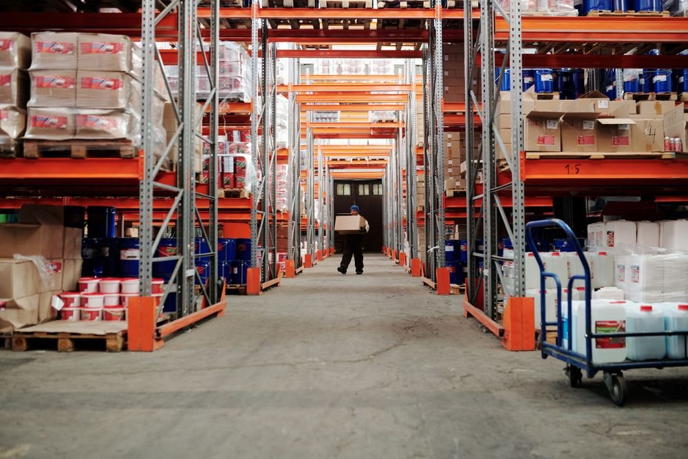 Worker carrying a box in an organized warehouse storage aisle, illustrating agentic commerce and logistics